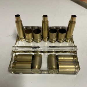 May include: A collection of brass bullet casings arranged in a clear, rectangular display case. The casings vary in size and are positioned upright and horizontally. The arrangement suggests a display or storage solution for ammunition components.
