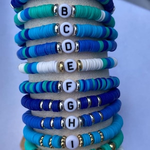 May include: Stack of ten bracelets with blue, white and turquoise beads. Each bracelet has a letter charm, starting with A and ending with K.