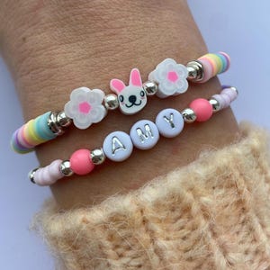 May include: Two bracelets with colourful beads, white flower charms, a bunny charm, and white letter beads spelling "AMY".