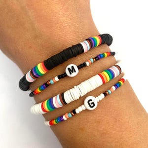 May include: A selection of colourful beaded bracelets. One black bracelet features rainbow-coloured accents. Two white bracelets and one black bracelet have the letters "M" and "G". The bracelets are made of small, flat, round beads.