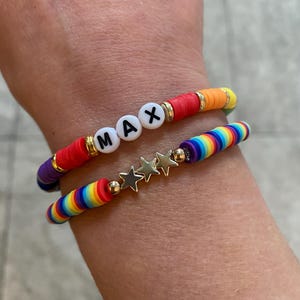 May include: Two bracelets on a wrist. The top bracelet is red, orange, and yellow with white beads that spell out "MAX". The bottom bracelet is rainbow coloured with gold star charms.