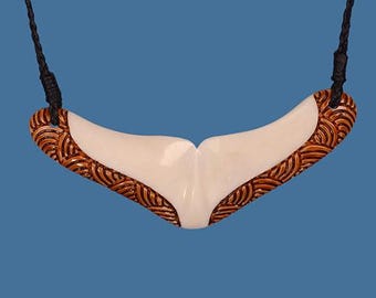 May include: A pendant necklace featuring a white, whale tail-shaped pendant with a brown, patterned border. The pendant hangs from a black cord against a blue background.