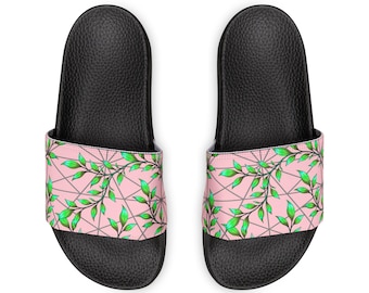 May include: A pair of black slide sandals featuring a wide strap with a light pink background, a dark gray geometric line pattern, and a vibrant green leaf vine design that wraps across the surface of the footwear.