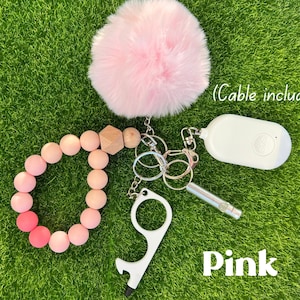 Può includere: Set portachiavi di sicurezza rosa con un soffice pompon rosa, un braccialetto di perline, un allarme bianco, un fischietto e uno strumento senza contatto. Il testo "Pink" è visibile. (Cavo incluso).