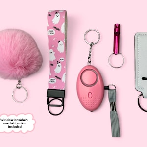 Könnte beinhalten: Eine Sammlung rosa Accessoires, darunter ein flauschiger Pompon-Schlüsselanhänger, eine pink-weißes Armband mit Geistermotiv, ein persönlicher Alarm in Pink, eine Pfeife und ein weißer Schlüsselanhänger. Die Artikel werden auf rosa Hintergrund präsentiert.