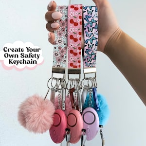 Puede incluir: Se muestran tres llaveros de seguridad con correas estampadas. Los estampados de las correas presentan flores, cerezas y mariposas. Cada llavero incluye una alarma personal, un pompón y un silbato. Texto: Crea tu propio llavero de seguridad.