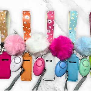 Puede incluir: Una colección de alarmas de seguridad personales con correas de muñeca a juego y pompones esponjosos. Las alarmas vienen en varios colores, incluyendo rosa, blanco, naranja, azul y verde. Las correas de muñeca presentan patrones florales y detalles brillantes.