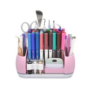 May include: A pink craft organizer filled with various tools and pens. The organizer has multiple compartments and holds scissors, pens, markers, and other crafting tools. The pens are in various colors, including blue, pink, and green.