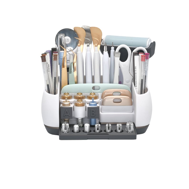 May include: A white and gray craft organizer filled with various tools. The organizer includes pens, tweezers, spatulas, scissors, a roller, and other crafting essentials. The tools are arranged in compartments for easy access and organization.