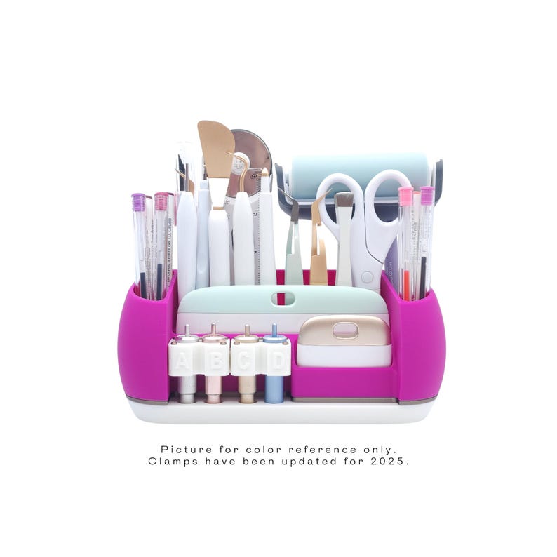 May include: A pink and white craft organizer filled with various tools, including scissors, pens, and a roller. The organizer has a mint green top and a white base. Text at the bottom reads "Picture for color reference only. Clamps have been updated for 2025."