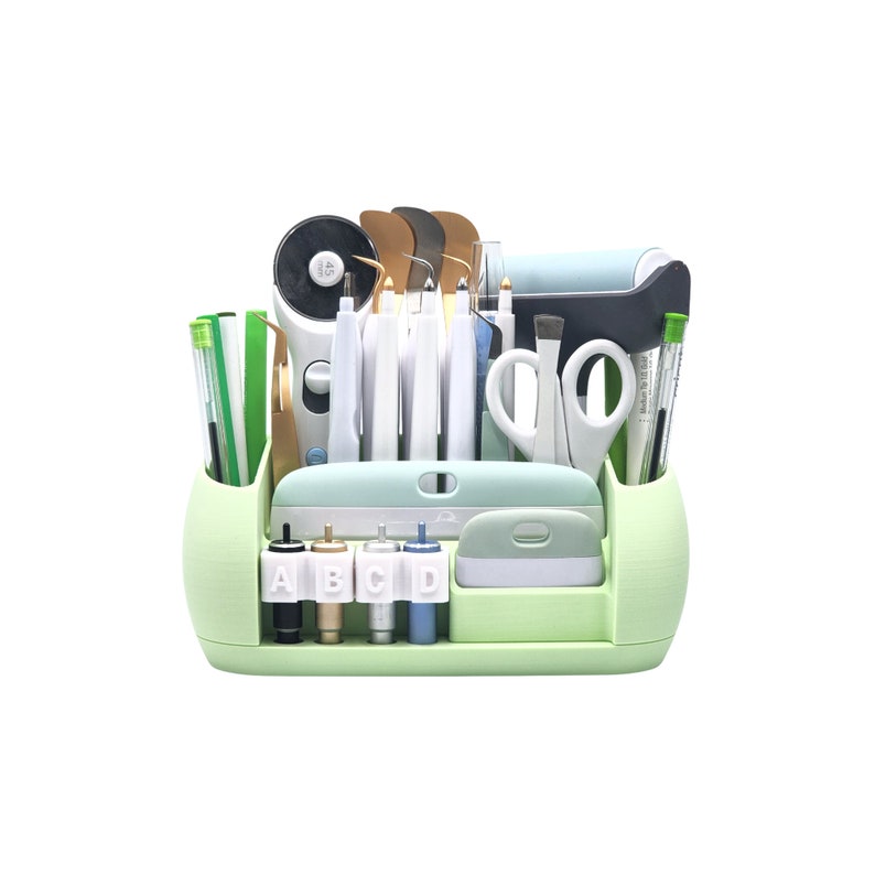 Puede incluir: Un organizador de manualidades verde claro lleno de varias herramientas, incluyendo tijeras, bol&iacute;grafos y cuchillas. El organizador tiene m&uacute;ltiples compartimentos y una peque&ntilde;a bandeja. Las herramientas est&aacute;n dispuestas ordenadamente, sugiriendo un enfoque en la organizaci&oacute;n y la artesan&iacute;a.