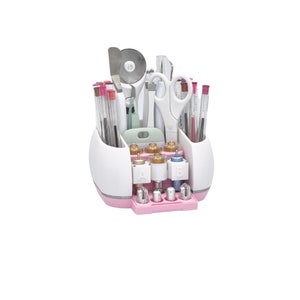 Puede incluir: Un organizador de manualidades blanco y rosa lleno de varias herramientas, incluyendo tijeras, bol&iacute;grafos y un cortador rotatorio. El organizador tiene m&uacute;ltiples compartimentos y una base rosa, ideal para guardar suministros de manualidades.