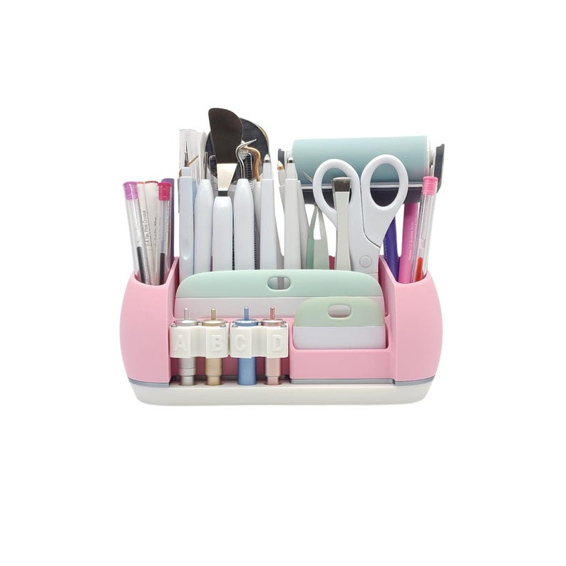 May include: A pink and white craft organizer filled with various tools. The organizer includes pens, scissors, a roller, and other crafting tools. The organizer has a light green section and a section with the letters A, B, C, and D.