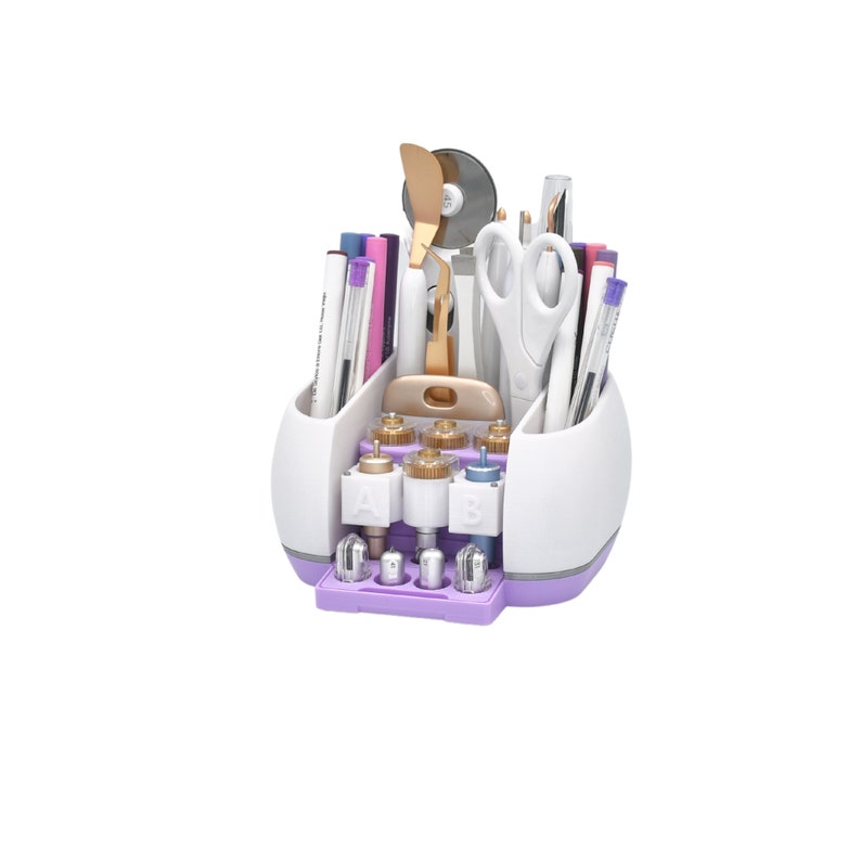 Puede incluir: Un organizador de manualidades blanco y morado lleno de varias herramientas, incluyendo bol&iacute;grafos, tijeras y una esp&aacute;tula. El organizador tiene m&uacute;ltiples compartimentos y una base con componentes etiquetados. Las herramientas est&aacute;n dispuestas para un f&aacute;cil acceso.