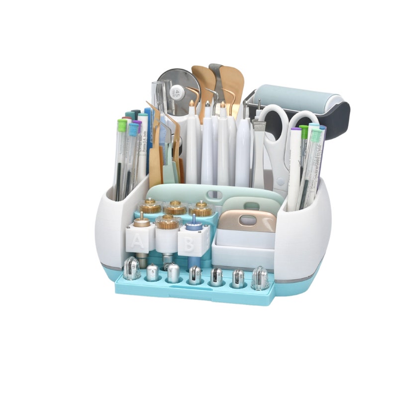May include: A white and light blue craft tool organizer filled with various tools, including pens, scissors, blades, and burnishing tools. The organizer has multiple compartments and a roller. The tools are arranged neatly.