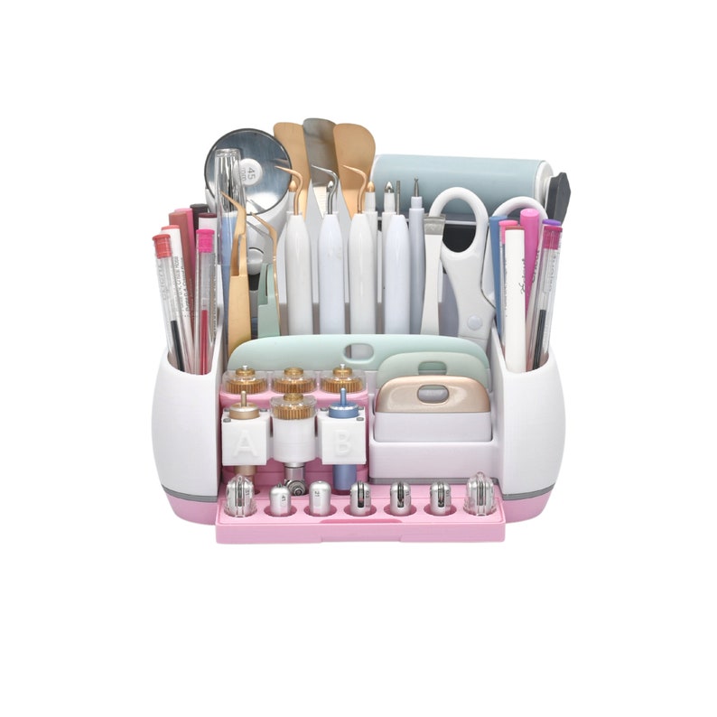 May include: A pink and white craft tool organizer filled with various tools. The organizer includes pens, scissors, spatulas, and cutting tools. The tools are arranged in a tiered design, with a variety of colors and shapes.