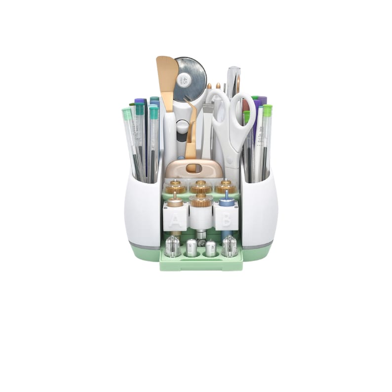 Puede incluir: Un organizador de manualidades blanco y verde lleno de varias herramientas, incluyendo bol&iacute;grafos, tijeras, una esp&aacute;tula y un cortador rotatorio. El organizador tiene m&uacute;ltiples compartimentos y una bandeja extra&iacute;ble, ideal para guardar suministros de manualidades.
