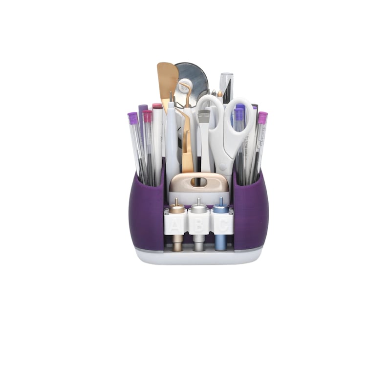 May include: A purple and white craft organizer filled with various tools. The organizer has two side compartments and a front section with three labeled slots. Tools include scissors, pens, and a spatula.
