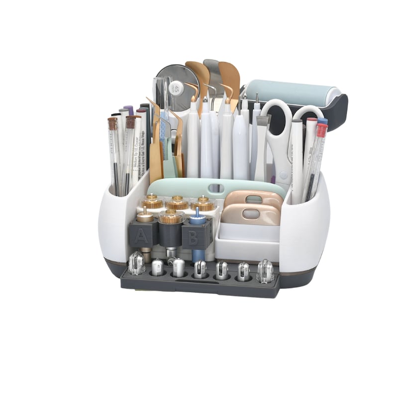 May include: A white and gray craft organizer filled with various tools, including pens, scissors, spatulas, and cutting blades. The organizer has multiple compartments and a dark gray base, ideal for crafting and DIY projects.