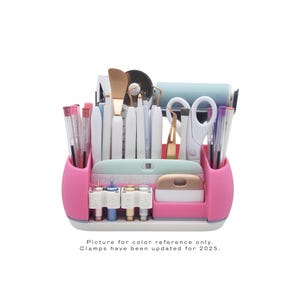 May include: A pink and white craft organizer filled with various tools, including scissors, pens, spatulas, and cutting blades. The organizer has multiple compartments and a light blue tray. Text at the bottom reads: "Picture for color reference only. Clamps have been updated for 2025."