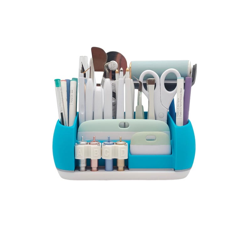May include: A teal and white craft organizer filled with various tools. The organizer has two teal side compartments holding pens and a purple pen. The center section holds white tools, scissors, and a light blue roller. The bottom has four color-coded tips.
