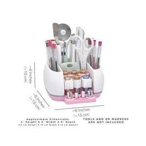 Puede incluir: Un organizador de manualidades blanco y rosa con varias herramientas y rotuladores. El organizador mide aproximadamente 10 cm de alto, 15 cm de ancho y 15 cm de profundidad. El texto de la imagen dice "TOOLS AND OR MARKERS ARE NOT INCLUDED."