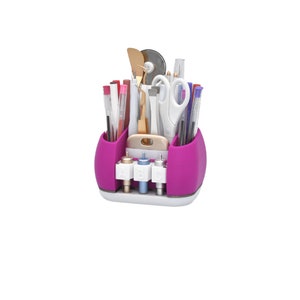 May include: A magenta craft organizer filled with various tools, including pens, scissors, tweezers, and a rotary cutter. The organizer has a white base and three labeled compartments at the bottom. The tools are arranged for easy access.