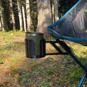 May include: A blue camping chair with a black cup holder attached to the armrest. The chair is set up in a forest setting.