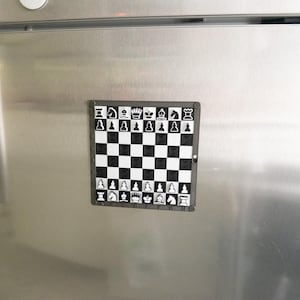 May include: A black and white magnetic chessboard with a silver frame. The chessboard is attached to a silver refrigerator.