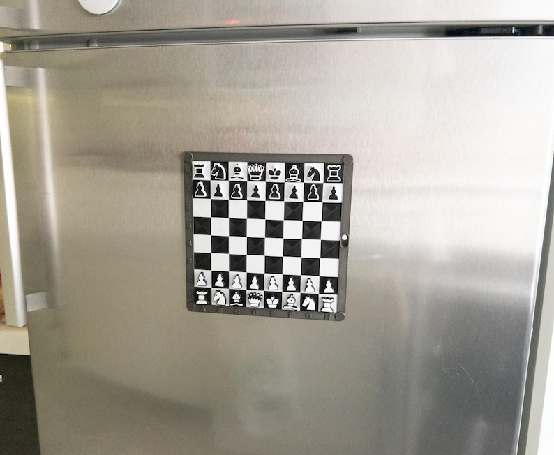 May include: A black and white magnetic chessboard with a silver frame. The chessboard is attached to a silver refrigerator.