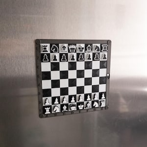 May include: A black and white magnetic chessboard with a black frame. The chessboard has a white background with black squares and black pieces. The pieces are arranged in the starting position for a game of chess.