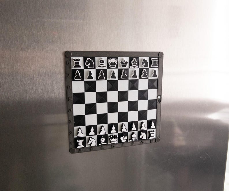 May include: A black and white magnetic chessboard with a black frame. The chessboard has a white background with black squares and black pieces. The pieces are arranged in the starting position for a game of chess.