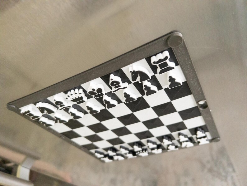 May include: A black and white magnetic chess set with all the pieces in place on a black and white checkered board. The board is attached to a black frame.