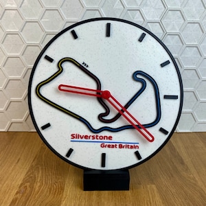 May include: A white clock with a black face and black hands. The clock face features a stylized outline of the Silverstone racetrack in yellow, blue, and red. The text "Silverstone Great Britain" is printed below the track.