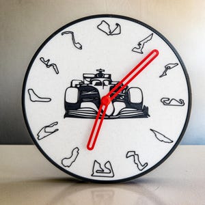 May include: A black and white clock with a red hand. The clock face features a silhouette of a race car and various race track layouts.