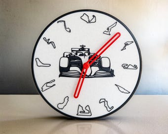 May include: A black and white clock with a red hand. The clock face features a silhouette of a race car and various race track layouts.