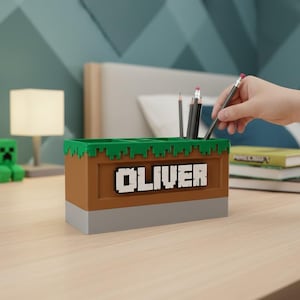 以下が含まれることがあります： Minecraftをテーマにした鉛筆立て。茶色で、緑色の上面と灰色のベースがあり、「OLIVER」という名前が白いピクセル化された文字で表示されています。鉛筆が中に入っています。シーンは木製の机の上にあります。