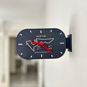 May include: A rectangular wall clock with rounded corners featuring a dark textured face, white hour markers, and bright red hands. The centre displays the Austin race track outline, the text AUSTIN, UNITED STATES GRAND PRIX, 2012, 5.513km, and 56.