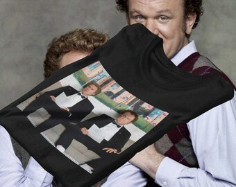 Op de afbeelding: Zwart T-shirt met een bedrukte afbeelding van twee mannen in zwarte pakken en vlinderdas die op een bank zitten. De mannen lachen en houden papieren vast. De achtergrond is een wazige afbeelding van een gebouw en bomen.