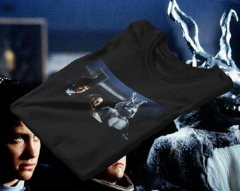 Op de afbeelding: Zwart t-shirt met een filmstill met twee mensen en een konijnachtig figuur. Het ontwerp is een filmreferentie. De achtergrond is donker, met een blauwe tint. Het t-shirt is gemaakt van zacht materiaal.