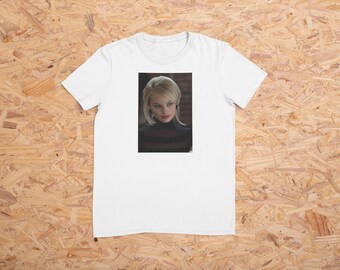 Op de afbeelding: Wit T-shirt met een portret van een vrouw met blond haar en een zwarte coltrui. De afbeelding is gecentreerd op het shirt, waardoor een eenvoudig maar opvallend ontwerp ontstaat. Het shirt is gemaakt van een zacht, comfortabel materiaal.
