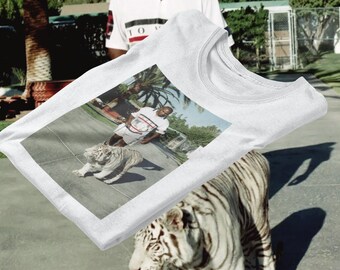 Op de afbeelding: Wit T-shirt met een bedrukte afbeelding van een man die een witte tijger uitlaat. De afbeelding is tegen een achtergrond van een met palmbomen beplante oprit.