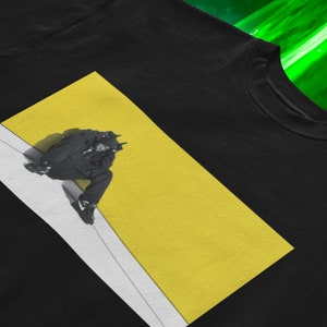 Può includere: T-shirt nero girocollo con un design grafico. Il design presenta un'immagine in bianco e nero di una persona seduta su uno sfondo giallo e bianco. La persona indossa un completo e scarpe da ginnastica nere.