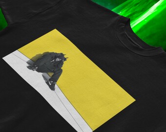 Op de afbeelding: Zwart T-shirt met ronde hals en een grafisch ontwerp. Het ontwerp toont een zwart-wit afbeelding van een persoon die op een gele en witte achtergrond zit. De persoon draagt een zwarte outfit en sneakers.