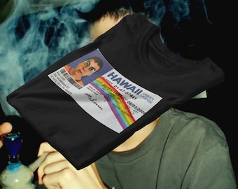 Op de afbeelding: Zwart T-shirt met een parodie op een Hawaïaanse rijbewijs. Het rijbewijs toont de naam "McLovin" en een foto. Het shirt heeft een regenboogontwerp en de tekst "HAWAII DRIVER LICENSE".
