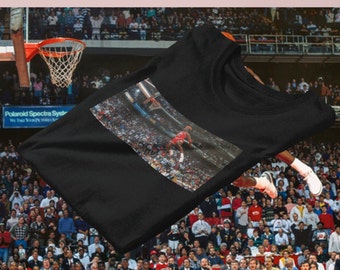 Op de afbeelding: Zwart T-shirt met ronde hals en een foto van een basketballer in de lucht, op het punt om de bal te dunken. De afbeelding is geplaatst tegen de achtergrond van een vol stadion. Het shirt is een klassieke, casual stijl.