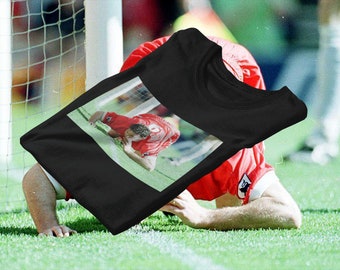 Op de afbeelding: Zwart T-shirt met een foto van een voetballer in een rood shirt op een groen veld. De speler bevindt zich in de buurt van een doel en lijkt bedroefd. Het shirt heeft een klassieke stijl.