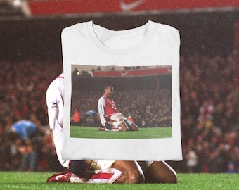 Op de afbeelding: Wit t-shirt met een foto van een voetballer die op een groen veld knielt en een doelpunt viert. De speler draagt een rood-wit tenue. De achtergrond toont een stadion vol toeschouwers.