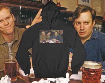 Op de afbeelding: Zwarte hoodie met een afbeelding van twee mannen in een pub. De afbeelding toont een man die zijn hand opsteekt. De achtergrond toont twee mannen, bierglazen en een pubinterieur.