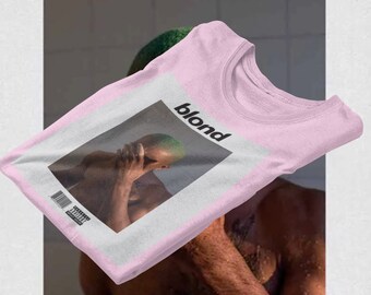 Op de afbeelding: Lichtroze t-shirt met de albumhoes van "Blond" van Frank Ocean. De afbeelding toont een persoon met groen haar en het woord "blond" in zwart. Het shirt heeft een ronde hals.